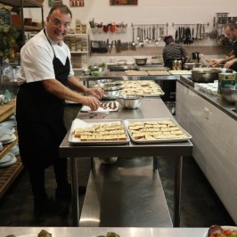 Smiling chef preparing food in a busy kitchen with trays of bread.