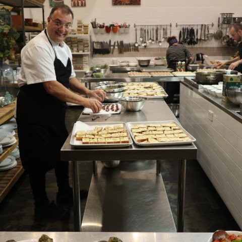 Smiling chef preparing food in a busy kitchen with trays of bread.