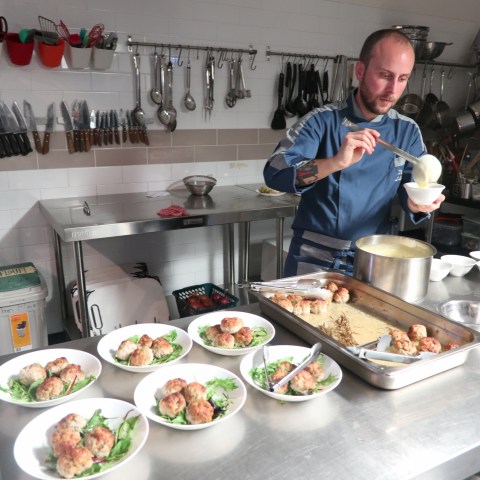 Chef preparing dishes with meatballs and greens in a commercial kitchen.