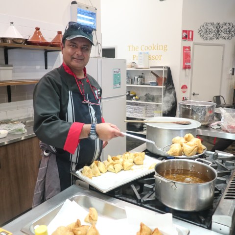 Chef frying samosas in a kitchen, with pots and tagine dishes visible.