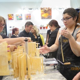 Group making pasta with flour, pasta machines, and drying racks in a cooking class setting.