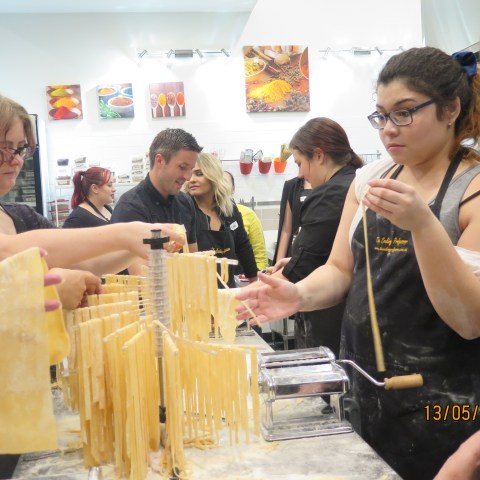 Group making pasta with flour, pasta machines, and drying racks in a cooking class setting.
