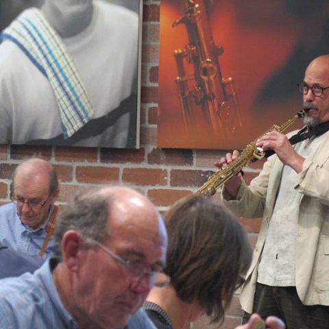 Man in a beige jacket playing saxophone in a restaurant with brick walls.