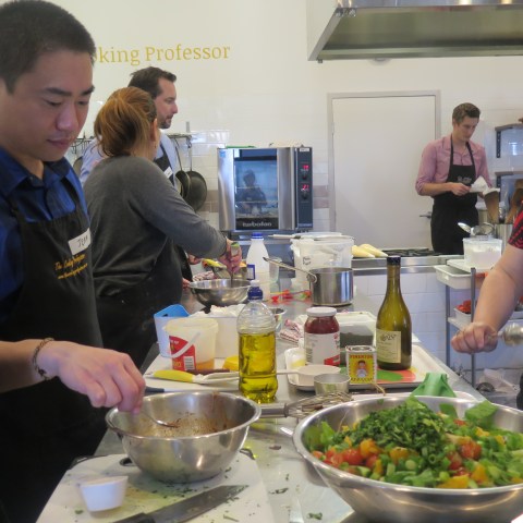 People in a kitchen classroom preparing food with various ingredients around them.