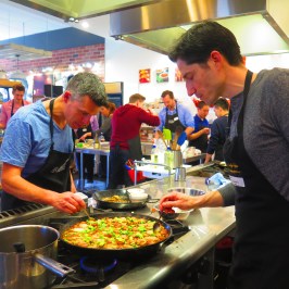 Two men cooking paella in a busy kitchen with others in the background.