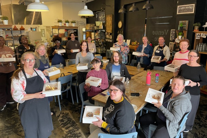 Group of people in a kitchen holding boxes, standing and seated at tables.