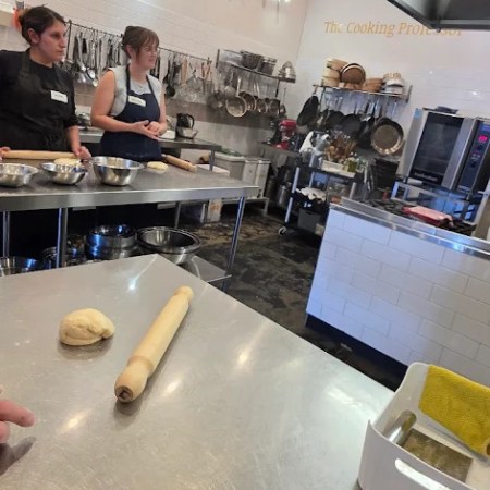 Two people in a kitchen with dough on a stainless steel counter.