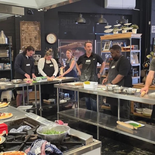 People in a kitchen preparing pizzas on metal tables.