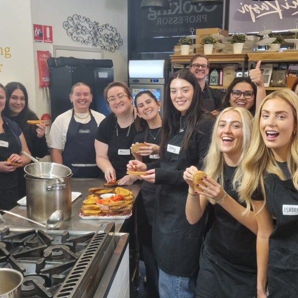 Group of people in aprons smiling and holding food in a kitchen setting.
