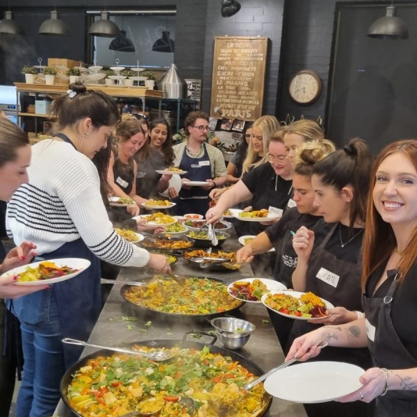 Group of people serving paella from large pans in a kitchen setting.